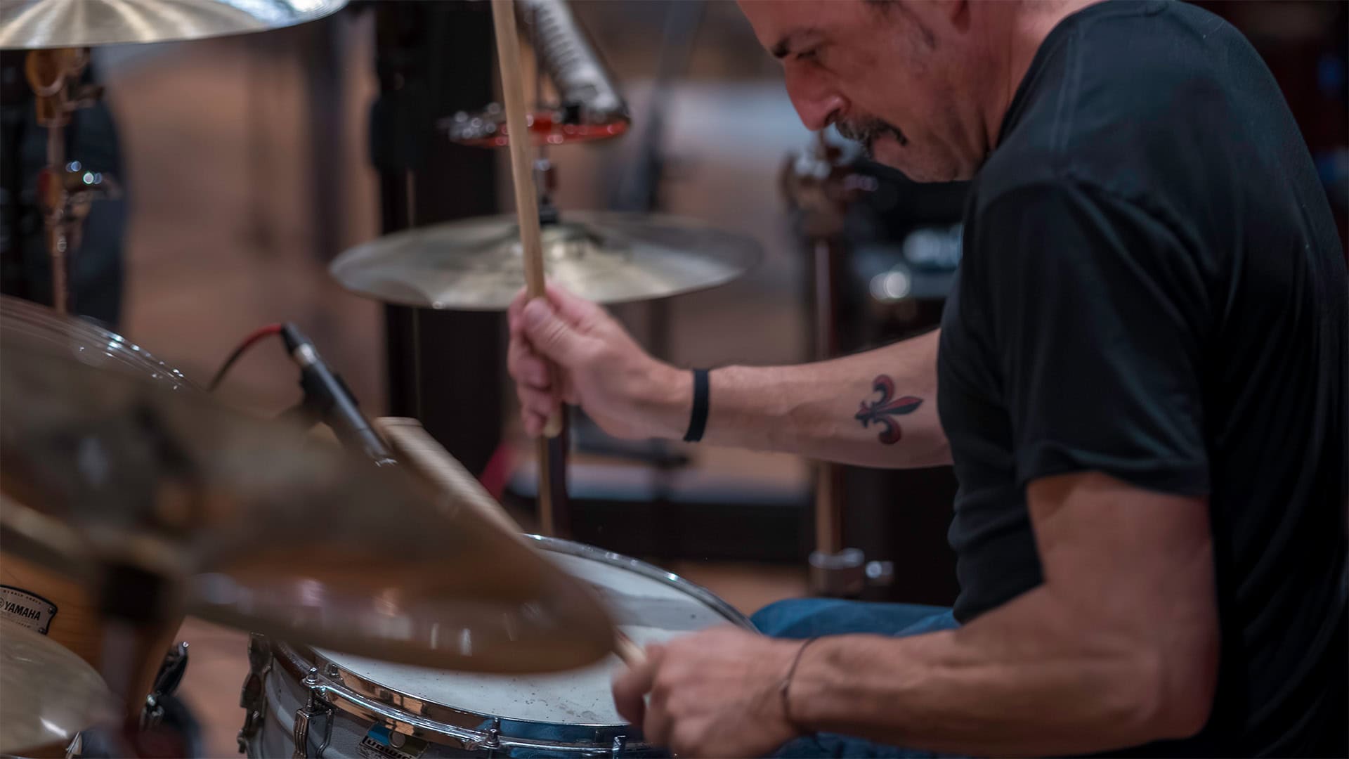 A man with a goatee plays the drums energetically, holding drumsticks mid-motion. He wears a black t-shirt and wristband, with a fleur-de-lis tattoo on his right arm—lost in music composing as the background blurs behind him.