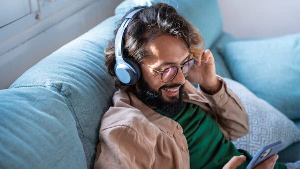 A man with glasses and headphones relaxes on a blue couch, smiling while looking at his smartphone.