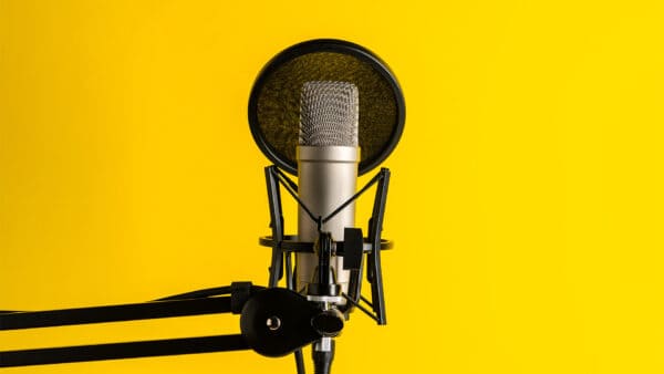 A silver studio microphone with a pop filter and adjustable arm stand is positioned in front of a bright yellow background.