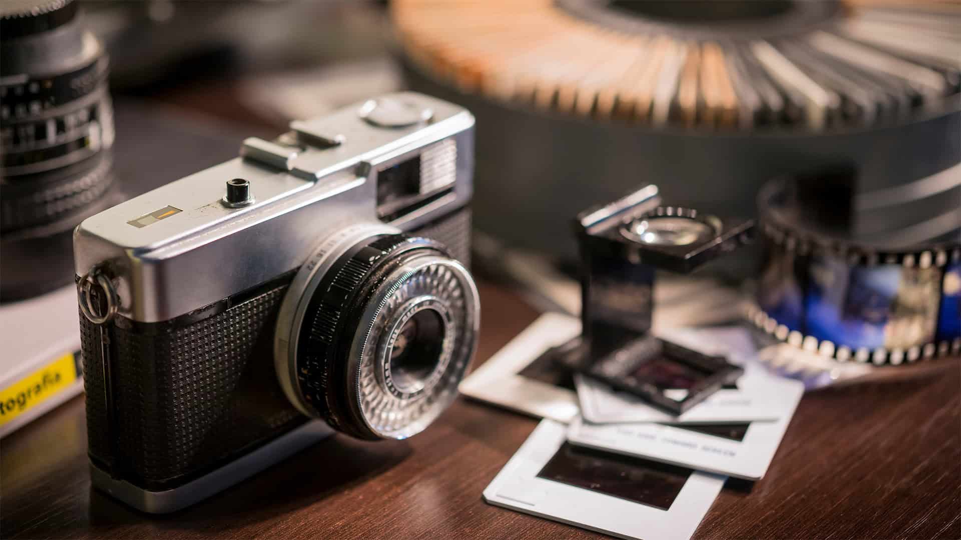A vintage camera sits on a table next to photo slides, a magnifier, a roll of film, and a circular slide holder, highlighting classic photography equipment perfect for photo scanning or photo digitization.