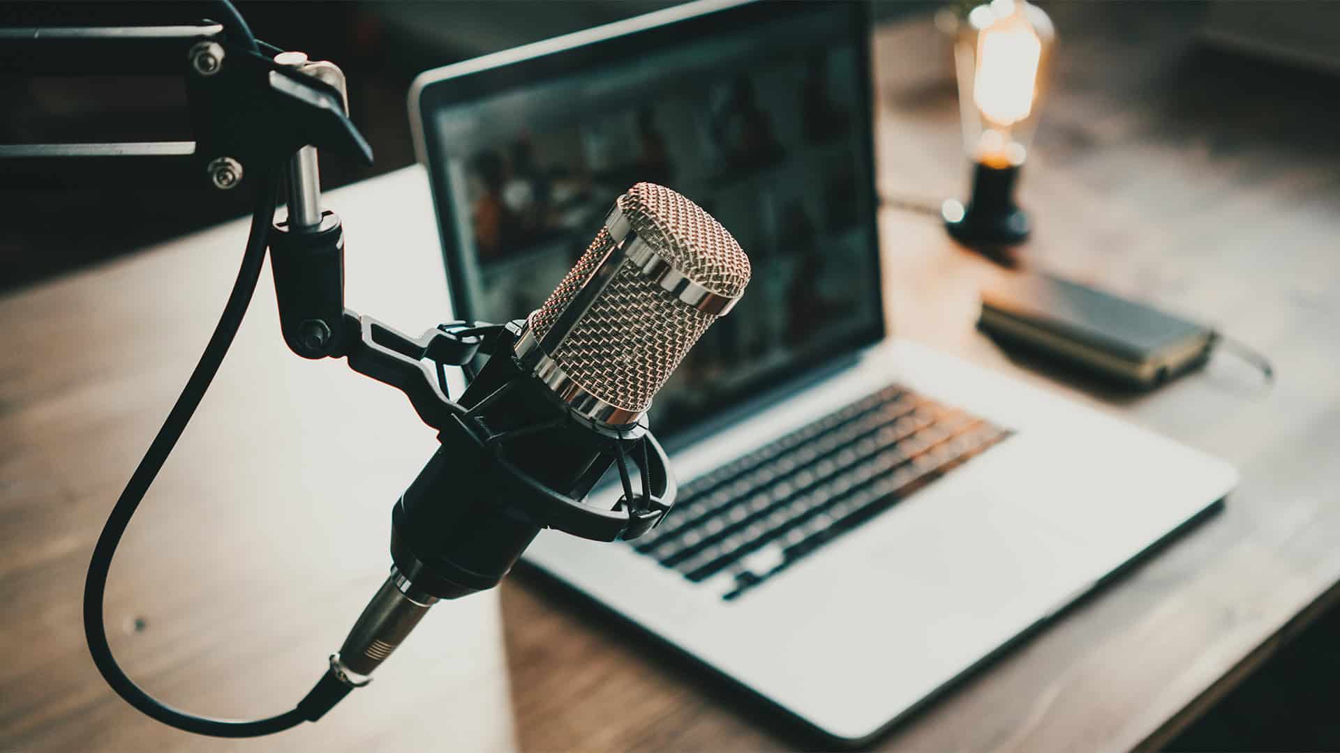 A professional microphone on a stand is positioned in front of an open laptop on a wooden desk, with a notebook and a lit lightbulb lamp in the background.