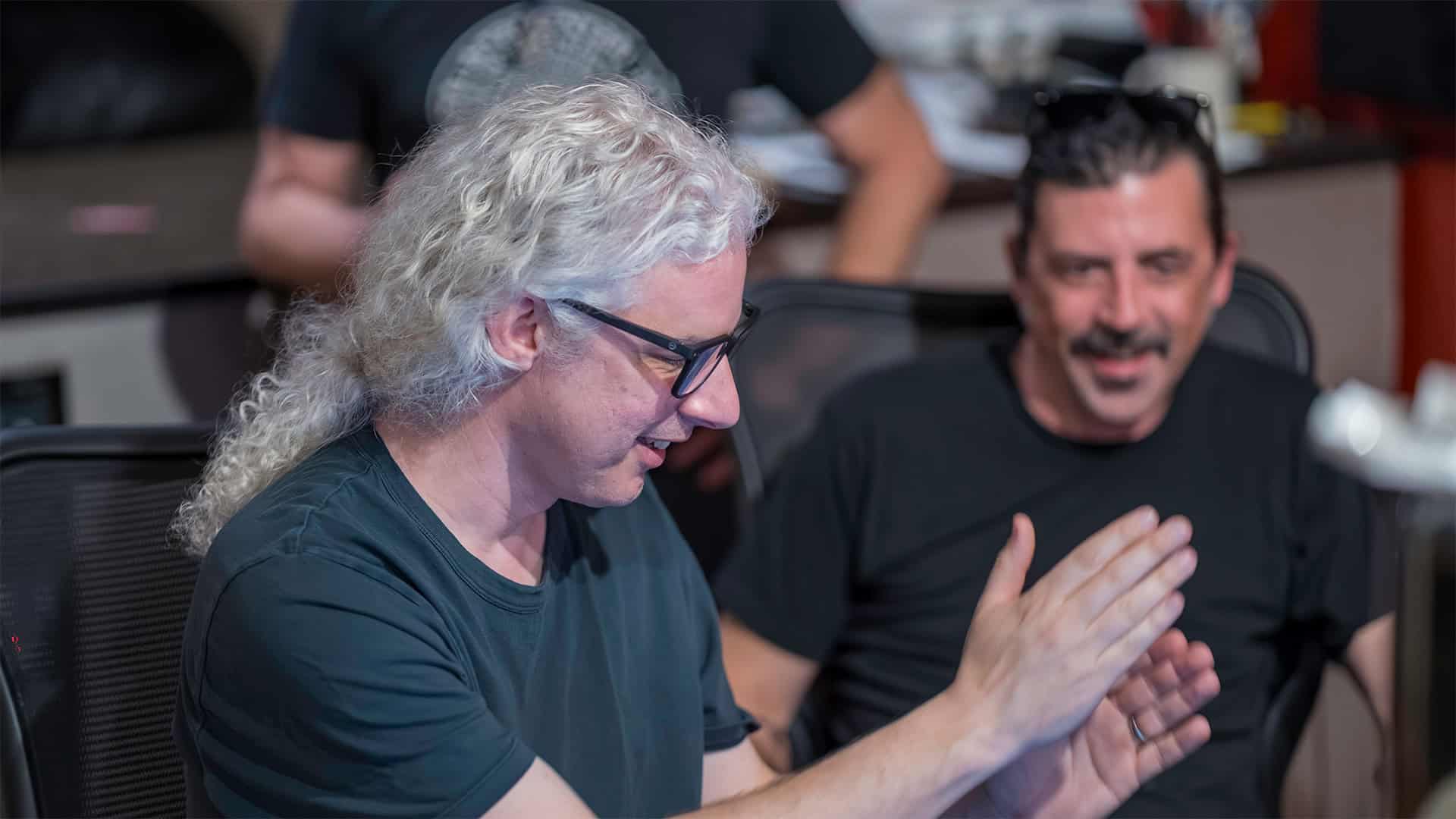 Two men sit indoors, both in black shirts. The man in the foreground, with long curly gray hair and glasses, claps his hands—possibly during audio recording training. Behind him, a dark-haired man with a mustache smiles.
