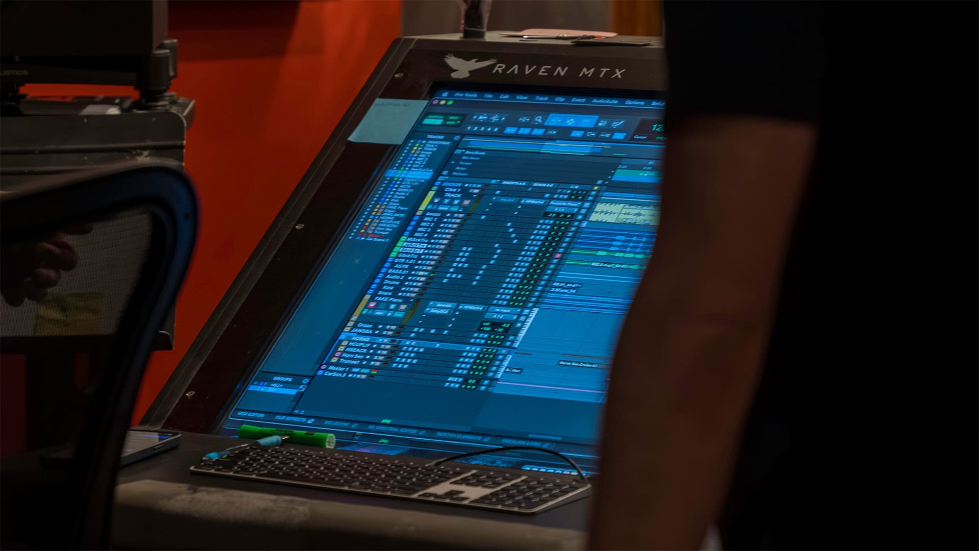 A large touchscreen computer labeled “Raven MTX” displays digital audio editing software and a music library. A keyboard and mouse sit below the screen, and a person stands nearby, partially visible on the right.