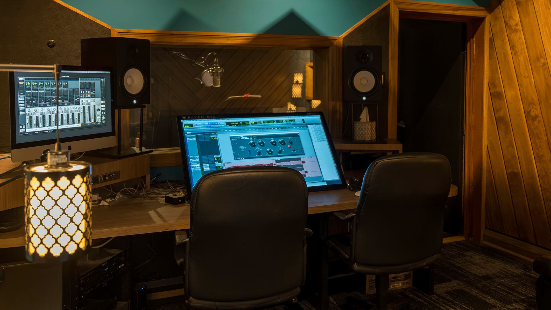 A professional recording studio with two chairs, a large computer monitor displaying audio editing software, studio speakers, a microphone in a sound booth, and warm ambient lighting.