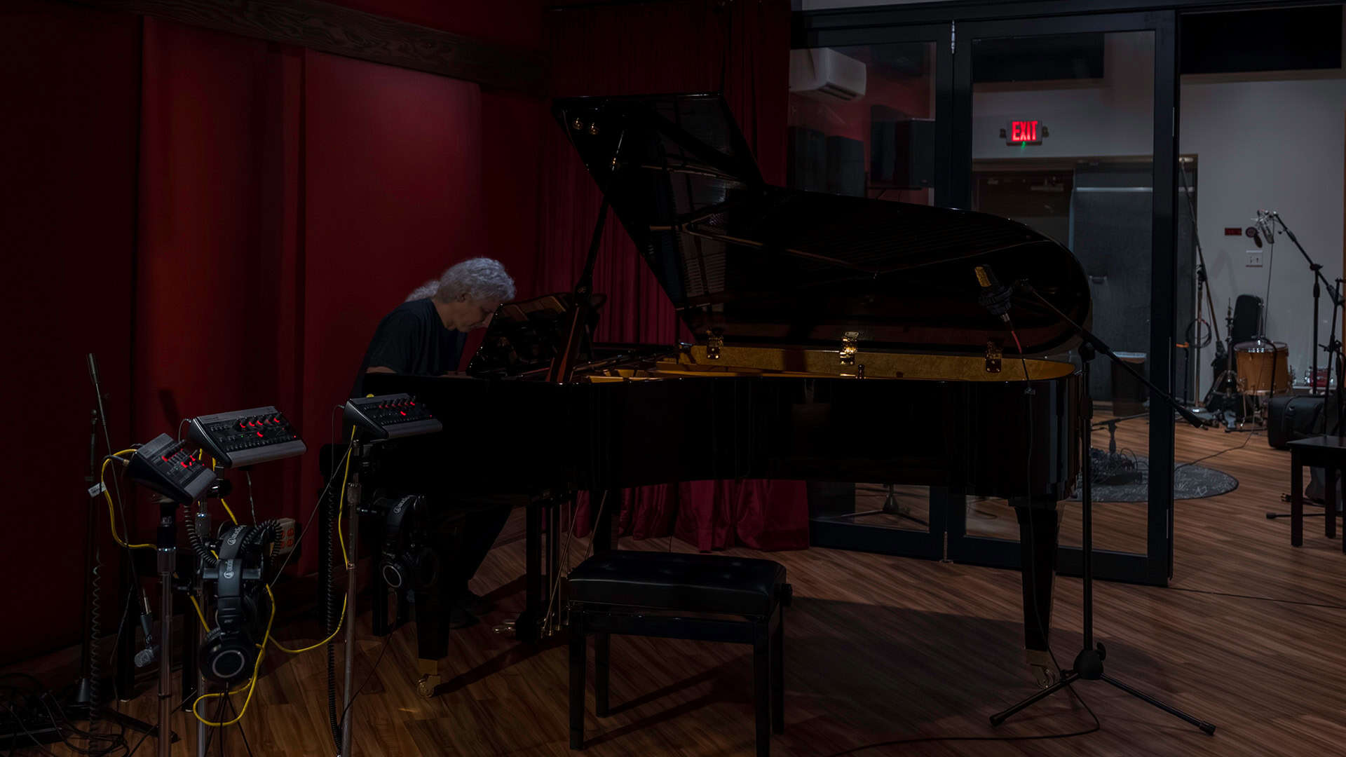A person with gray hair plays a grand piano in a dimly lit recording studio with red walls, wooden floor, and audio equipment nearby. A glass wall separates another room with instruments.