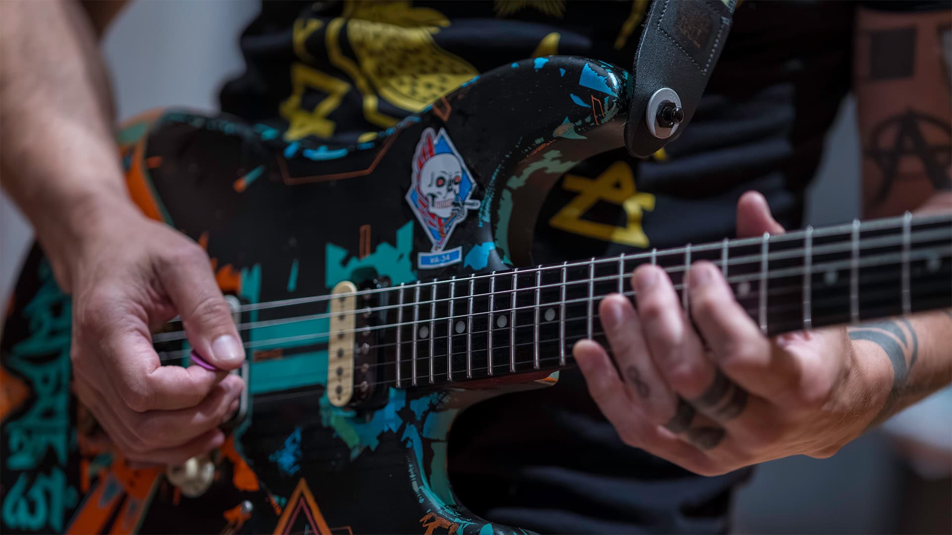 A person playing an electric guitar decorated with colorful stickers and artwork, their tattooed fingers pressing the strings on the fretboard as part of an audio recording training session.