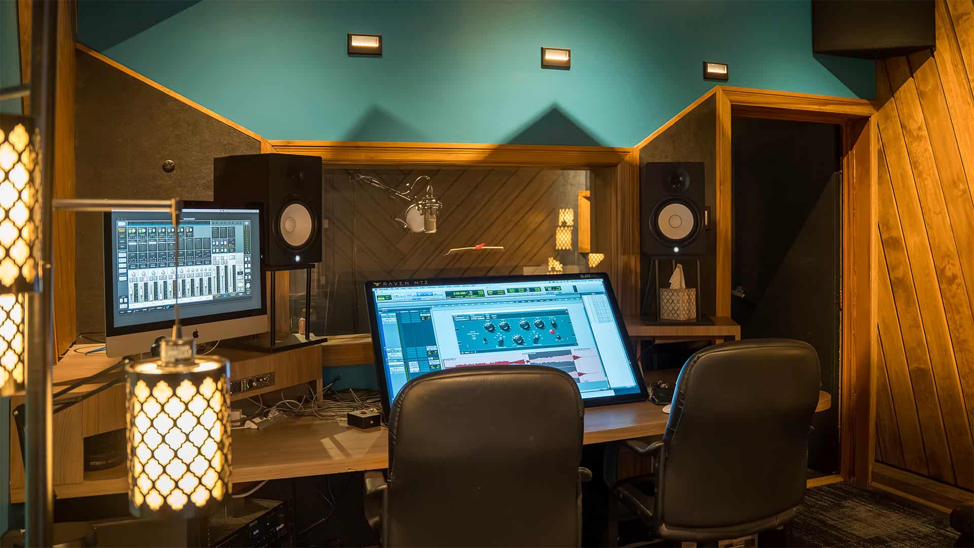 A modern recording studio designed for foreign language recording, featuring two chairs, a large computer monitor displaying audio editing software, wall-mounted speakers, a microphone, and warm lighting from hanging lamps.