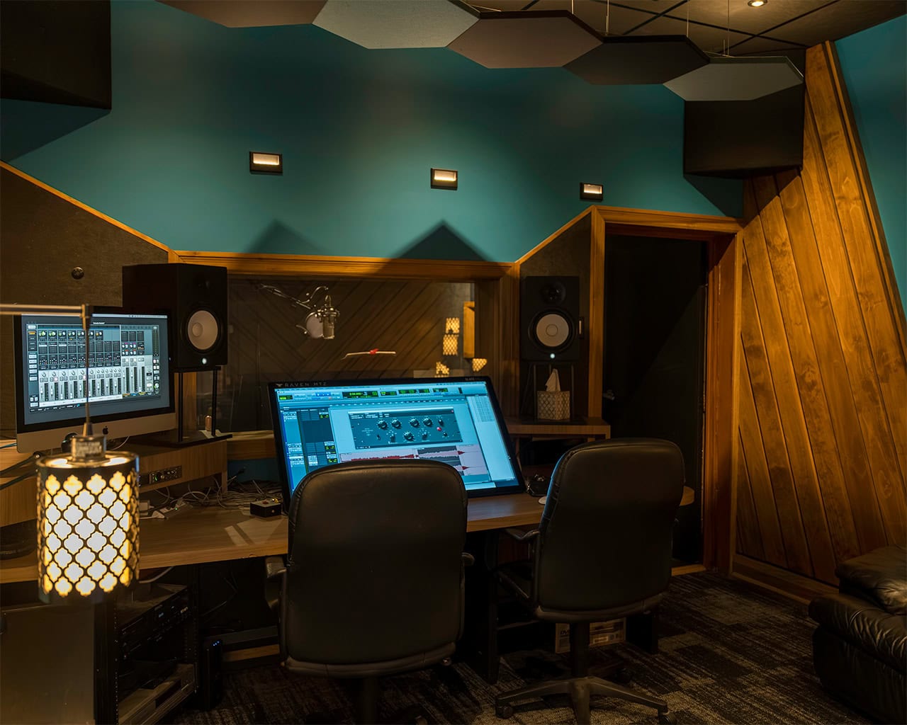 A recording studio with two chairs, computer monitors displaying audio software, speakers, a microphone, and wood-paneled walls under soft lighting. The atmosphere is cozy and professional.