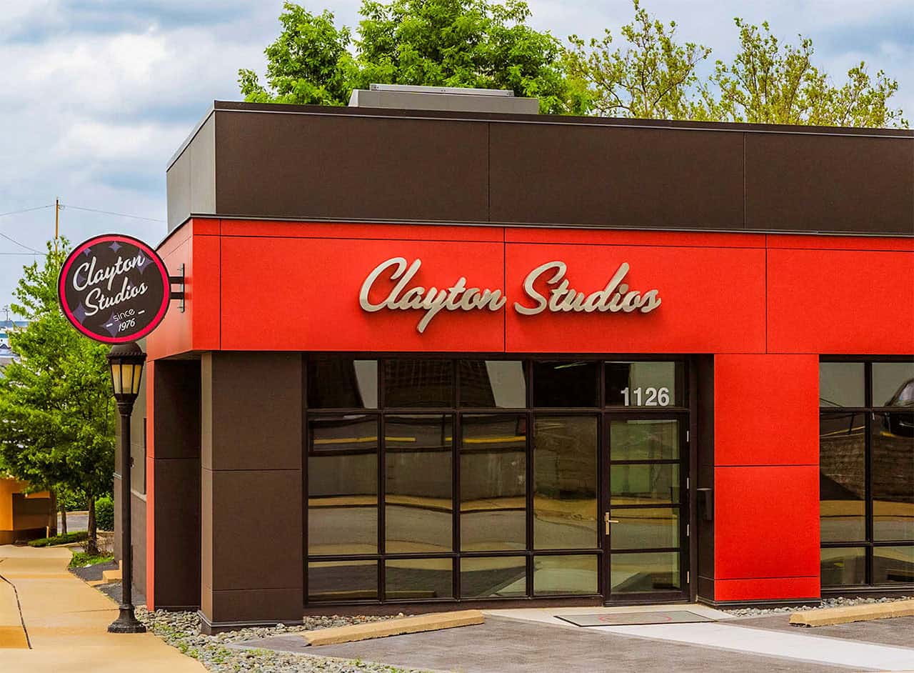 A modern building with a red and brown exterior displays a sign reading "Clayton Studios" above glass doors and windows, with a matching circular sign on a nearby post. The address "1126" is visible on the building.