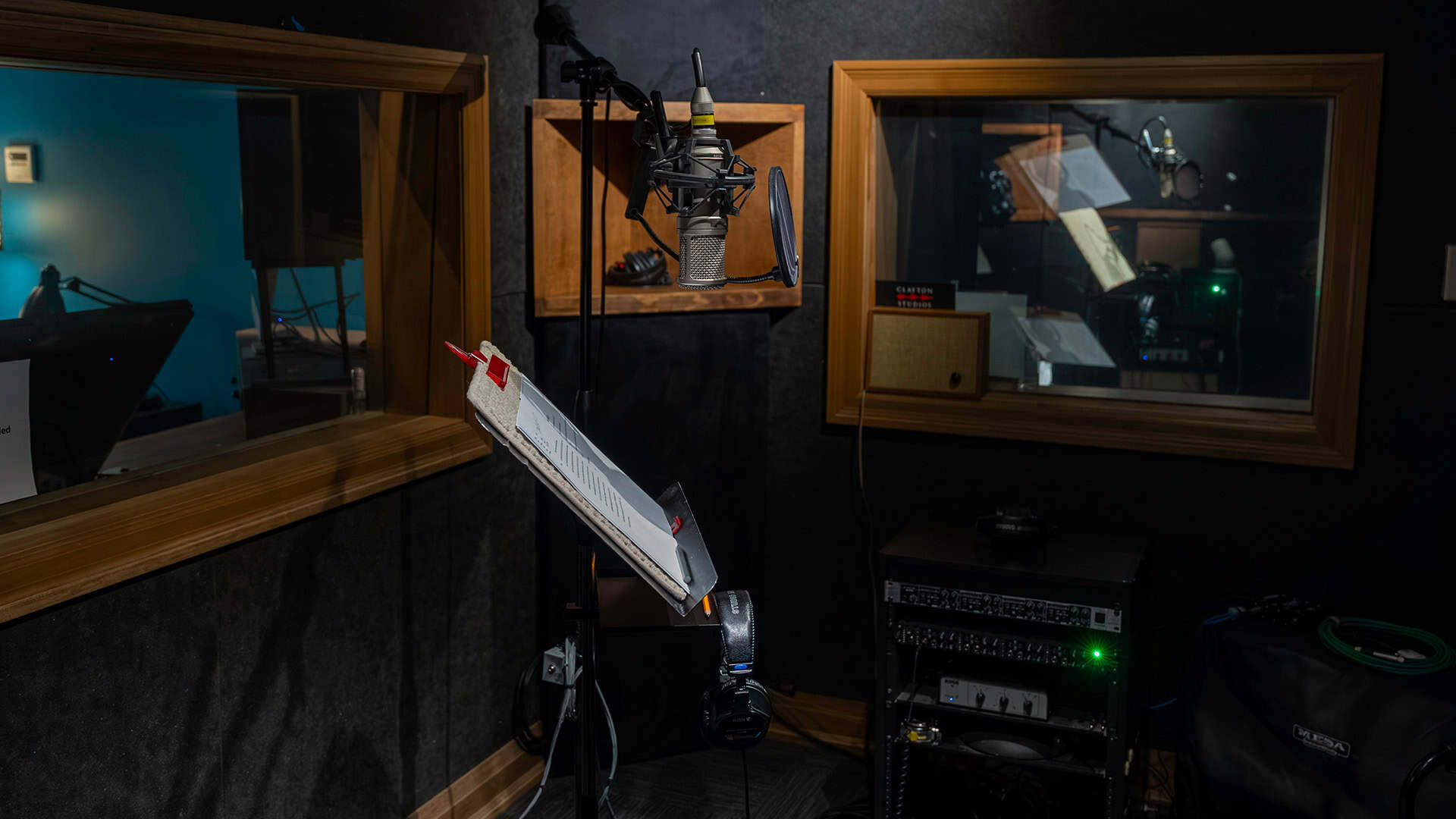 A recording studio with a microphone, headphones, and music stand holding a script. Control room windows look into adjacent rooms with additional audio equipment and microphones visible. The room is dimly lit.